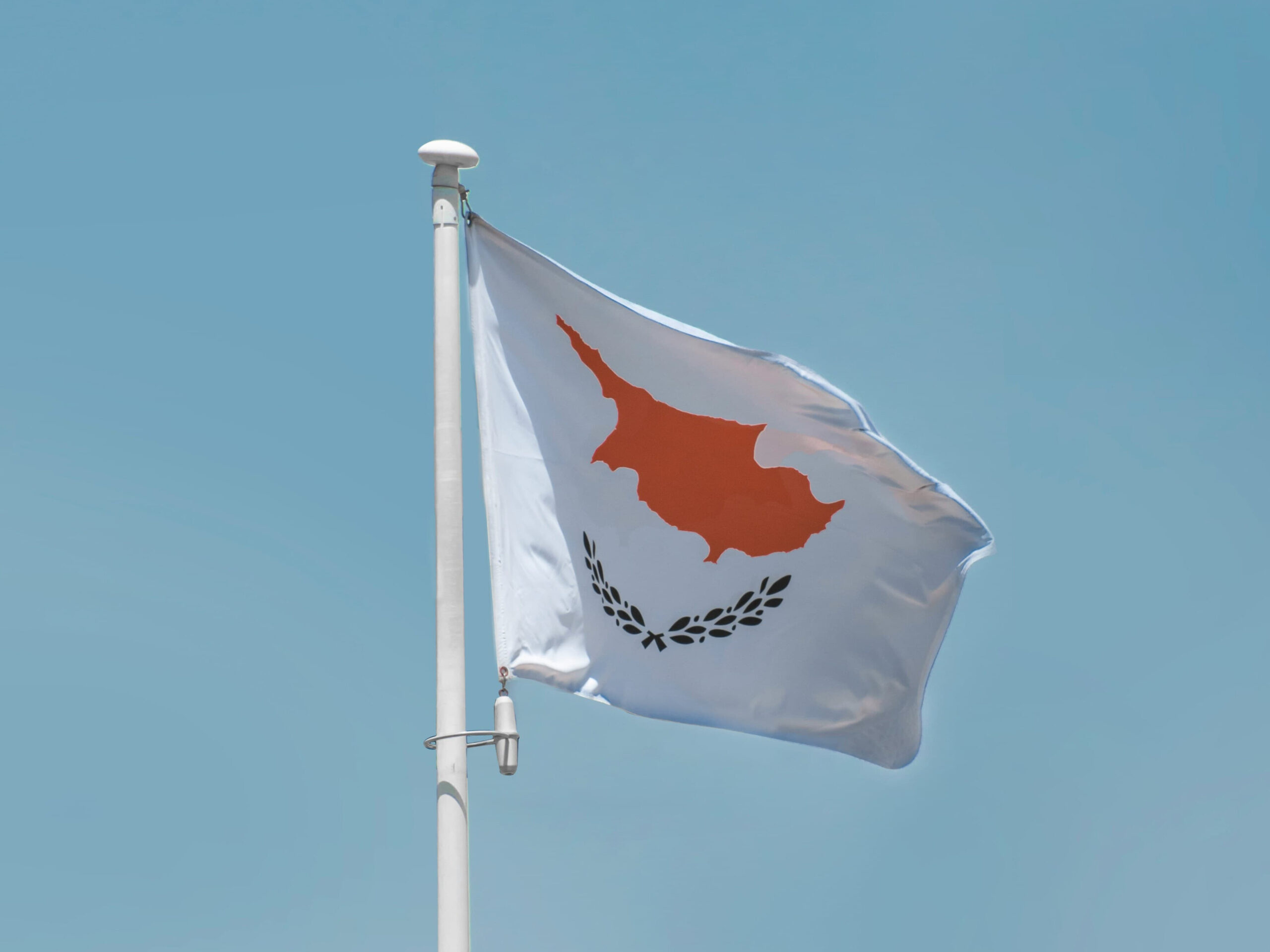 Cyprus national flag waving on a flagpole against a clear blue sky, featuring the orange island map and olive branches symbolizing peace.